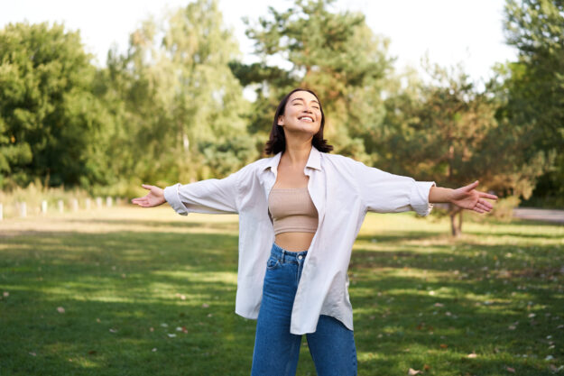 Carefree asian girl laughing and dancing in park, enjoying summer sunny day, raising hands up and breathing fresh air